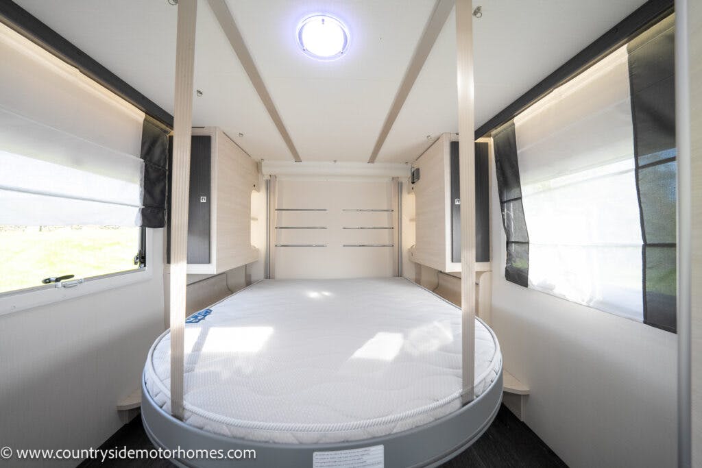Interior of the 2021 Chausson 778 Premium motorhome featuring a round, white mattress with light wooden accents and white walls. There are windows on either side with white and blackout blinds. The ceiling has a circular light source in the middle. Website URL is visible in the lower-left corner.