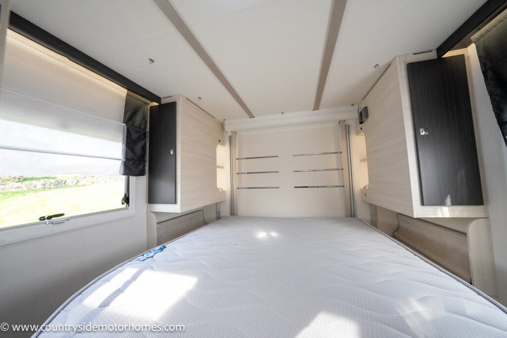 Interior of the 2021 Chausson 778 Premium motorhome featuring a neatly made bed with a white mattress, light wooden cabinetry on both sides, and windows with blinds partially drawn. The room is bright with natural light streaming in from the window on the left.