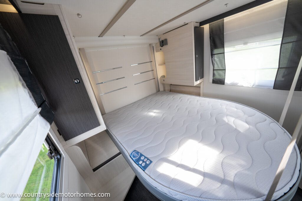 Interior of a 2021 Chausson 778 Premium motorhome bedroom featuring a neatly made bed with a rounded end, positioned on a platform. The room has modern decor with light wood finishes, dark accents, and large windows with block-out blinds. A website URL is visible on the lower-left corner.