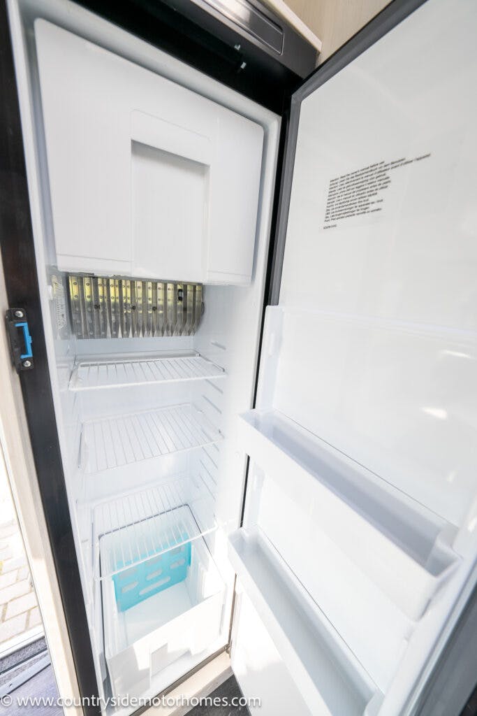 An open refrigerator with an empty freezer compartment at the top, several empty shelves in the main section, and two storage compartments on the door. A blue container is placed on the bottom shelf. The door bears the website www.countrysidemotorhomes.com, showcasing their 2021 Chausson 778 Premium.