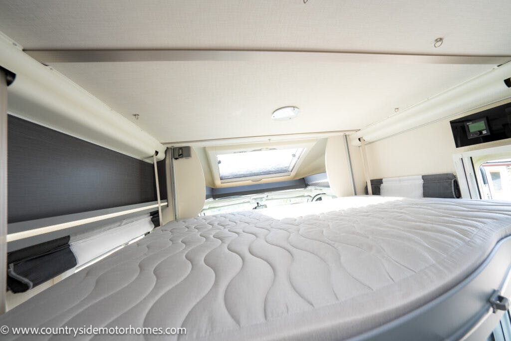 Interior view of a 2021 Chausson 778 Premium motorhome showing a neatly made bed with a textured grey mattress cover, overhead skylight, and side windows. Walls feature storage compartments and various fixtures. Website address www.countrysidemotorhomes.com is visible at the bottom left.