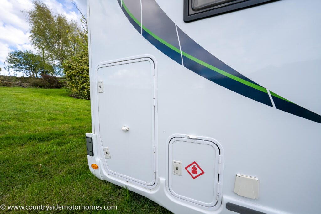 A close-up of the side of a 2021 Chausson 778 Premium motorhome with two compartment doors, one larger than the other. The motorhome sports a green and black stripe design. A warning symbol for flammable materials is on the smaller compartment door. Trees and grass are in the background.