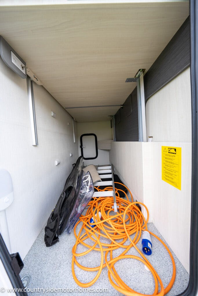 This image shows the interior storage compartment of a 2021 Chausson 778 Premium motorhome. The compartment contains an orange extension cord, a folded black bag, some plastic sheets, and other miscellaneous items. The space is enclosed with a door visible at the far end.