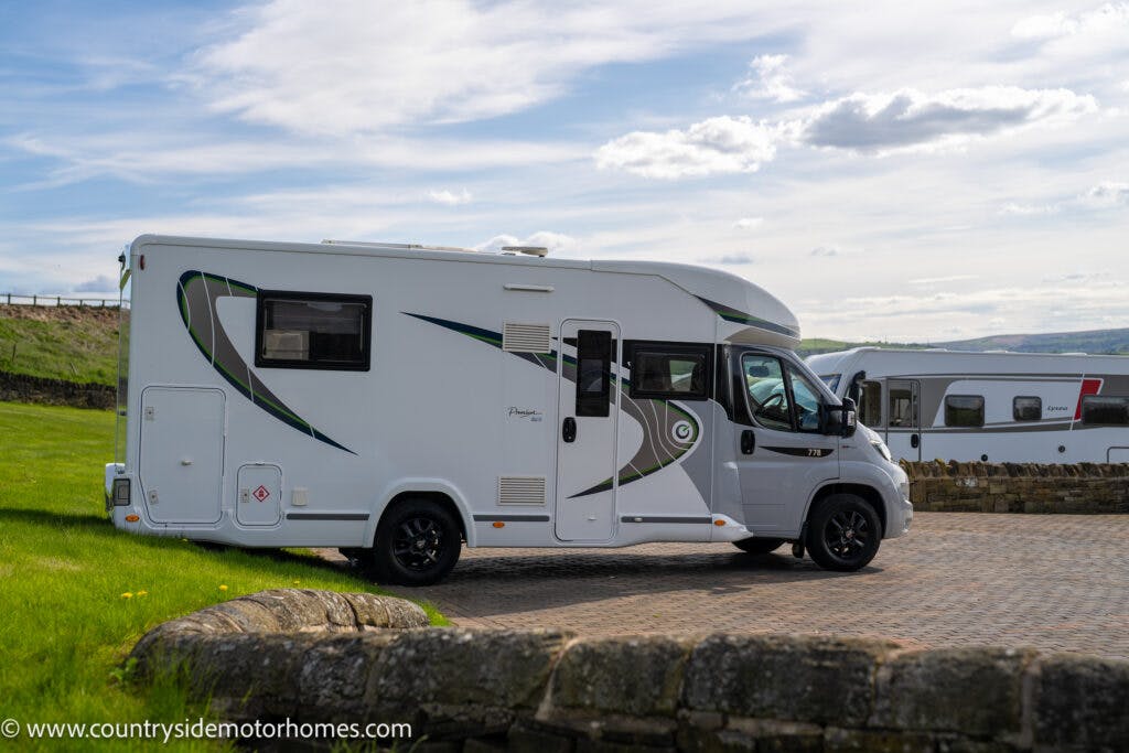 A 2021 Chausson 778 Premium motorhome with green and black accents is parked on a paved area near a stone wall. The website "www.countrysidemotorhomes.com" is visible in the bottom left corner. Another similar vehicle is in the background under a partly cloudy sky.