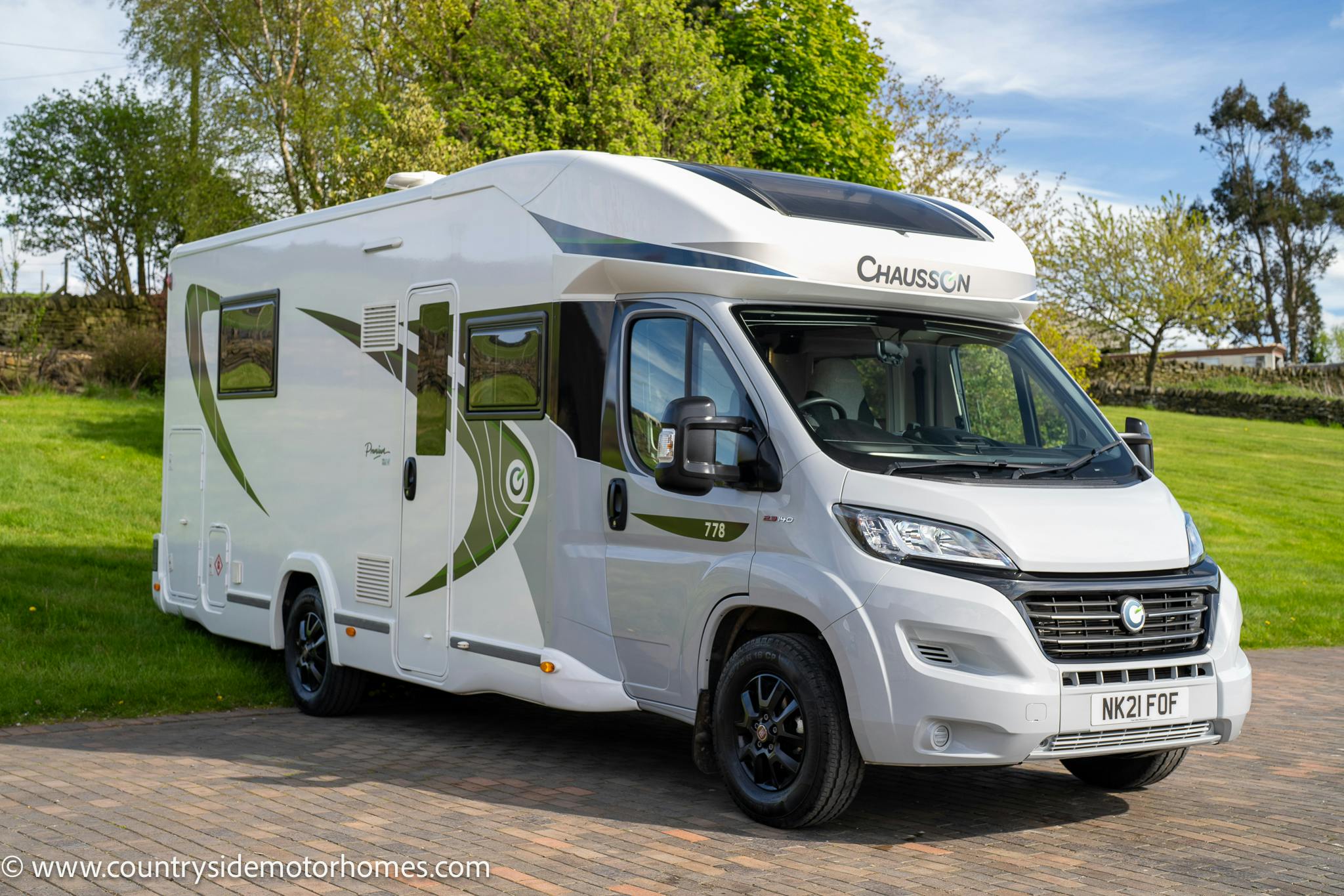 A white 2021 Chausson 778 Premium motorhome with green accents parked on a paved driveway. The model number "778" is displayed on the side, and the license plate reads "NX21 FDF." Trees and a grassy area are visible in the background. The website "www.countrysidemotorhomes.com" is noted.