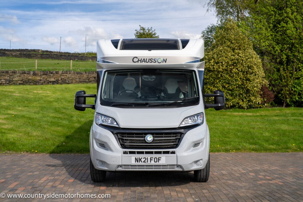 A 2021 Chausson 778 Premium motorhome is parked on a paved driveway with grass and trees in the background. The vehicle is white with the brand "Chausson" displayed prominently on the front. The license plate reads "NK21 FOF.