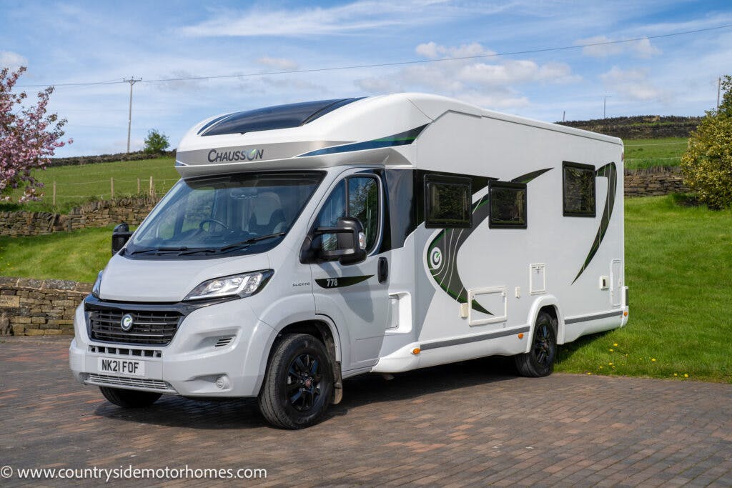 A white 2021 Chausson 778 Premium motorhome is parked on a stone driveway with green hills and a partly cloudy sky in the background. The motorhome has black and green accents and a visible registration plate reading "MX21 FDF". The image features a watermark reading "www.countrysidemotorhomes.com".