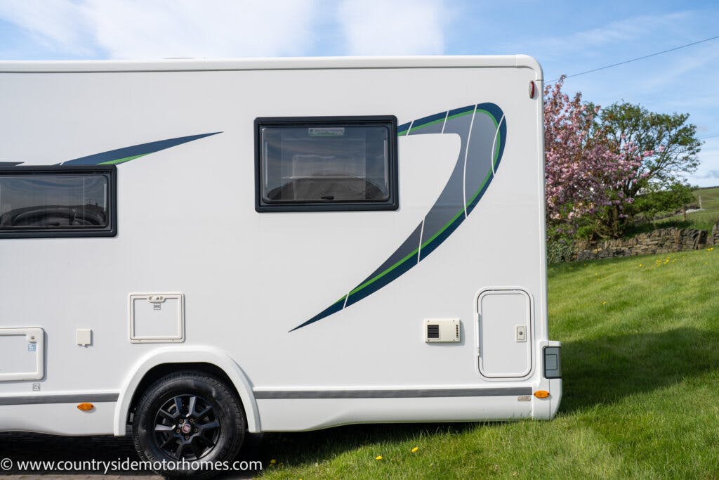 A side view of a 2021 Chausson 778 Premium motorhome with green and black decals, parked on a grassy area. The motorhome has one window and multiple storage compartments visible. There is a blooming tree and a stone fence in the background.