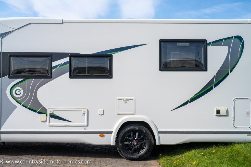 A side view of a white 2021 Chausson 778 Premium motorhome with black and green graphic designs. The motorhome features two windows, a small storage compartment, and a black wheel. The background shows a clear blue sky and some grass. The website "www.countrysidemotorhomes.com" is visible at the bottom left.