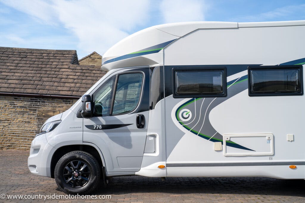 A white 2021 Chausson 778 Premium motorhome with black and green accents is parked on a brick driveway. The motorhome features windows along its side and branding that includes the number 778 and a logo. Roofed buildings are visible in the background.