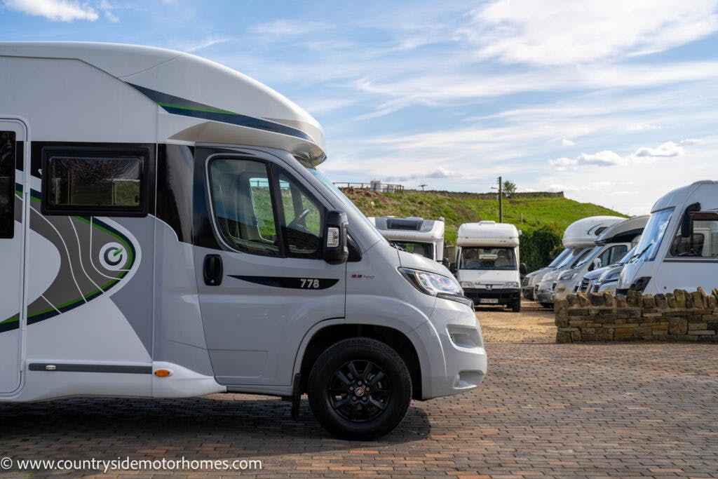 Several motorhomes are parked in an outdoor lot on a partly cloudy day. The 2021 Chausson 778 Premium in the foreground displays the number 778 and features a sleek white and gray design. The lot is paved with brick and surrounded by a short stone wall. URL visible: www.countrysidemotorhomes.com.