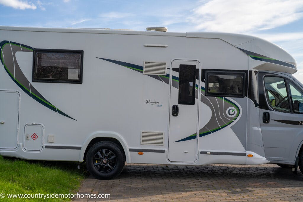 Side view of a white 2021 Chausson 778 Premium motorhome parked on a paved area next to a grassy patch. The motorhome has green and gray accents, multiple windows, and vents. The logo and website "countrysidemotorhomes.com" are visible on the side and front. The sky is partly cloudy.