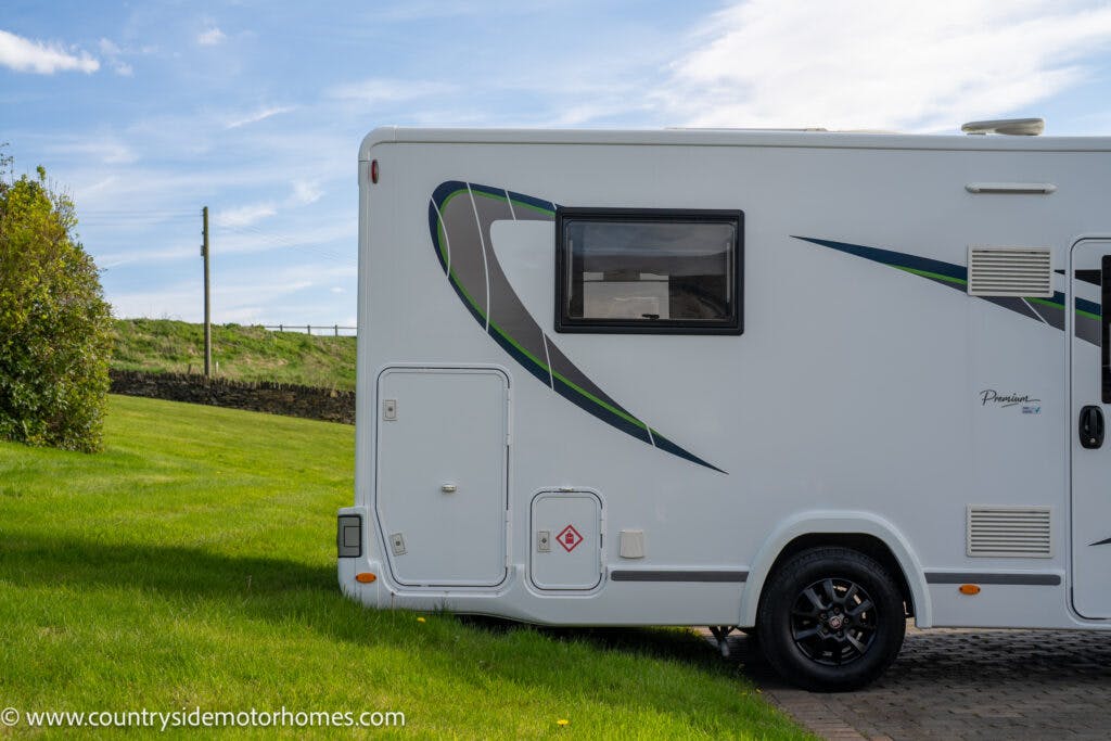 A white 2021 Chausson 778 Premium motorhome is parked on a paved surface next to a green grassy area. The side of the motorhome features a window, decorative green and black stripes, and several utility access panels. Blue sky and some wooden posts and a fence are visible in the background.