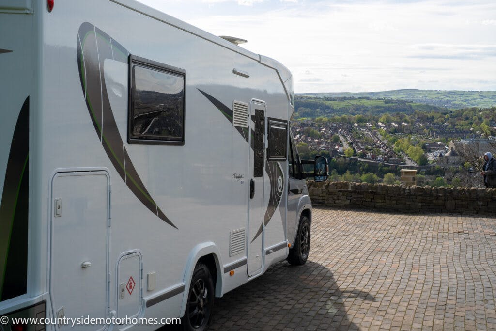 A white 2021 Chausson 778 Premium motorhome is parked on a paved area with a countryside view in the background. The vehicle has black and green striped designs on its side. A stone wall is visible nearby, with a person standing close to the edge. The website's URL, "www.countrysidemotorhomes.com," is seen in the bottom left corner.
