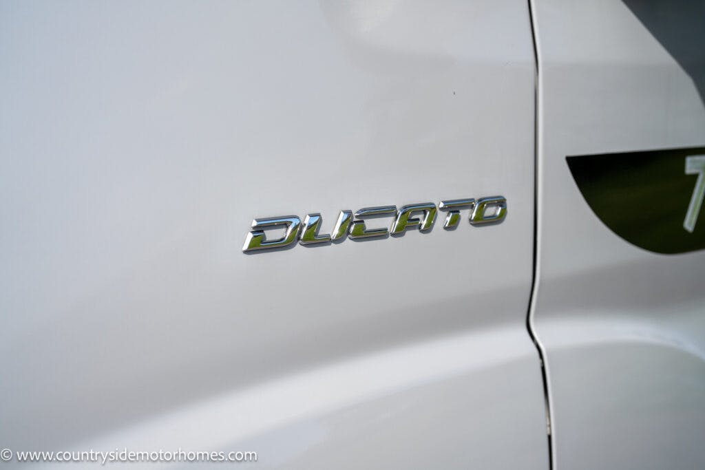 Close-up view of the "Ducato" emblem on the side of a white 2021 Chausson 778 Premium, showing the brand name in chrome lettering. The background includes part of the car door and a portion of a logo on the right side.