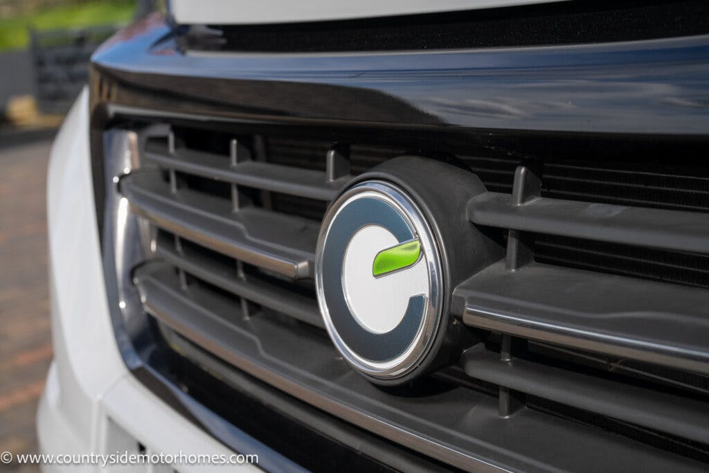 Close-up of a vehicle's front grille featuring a round emblem with a stylized green leaf design. The logo appears to be for Countryside Motorhomes, as indicated by the watermark in the bottom-left corner. This 2021 Chausson 778 Premium model showcases a predominantly black grille with some white elements.