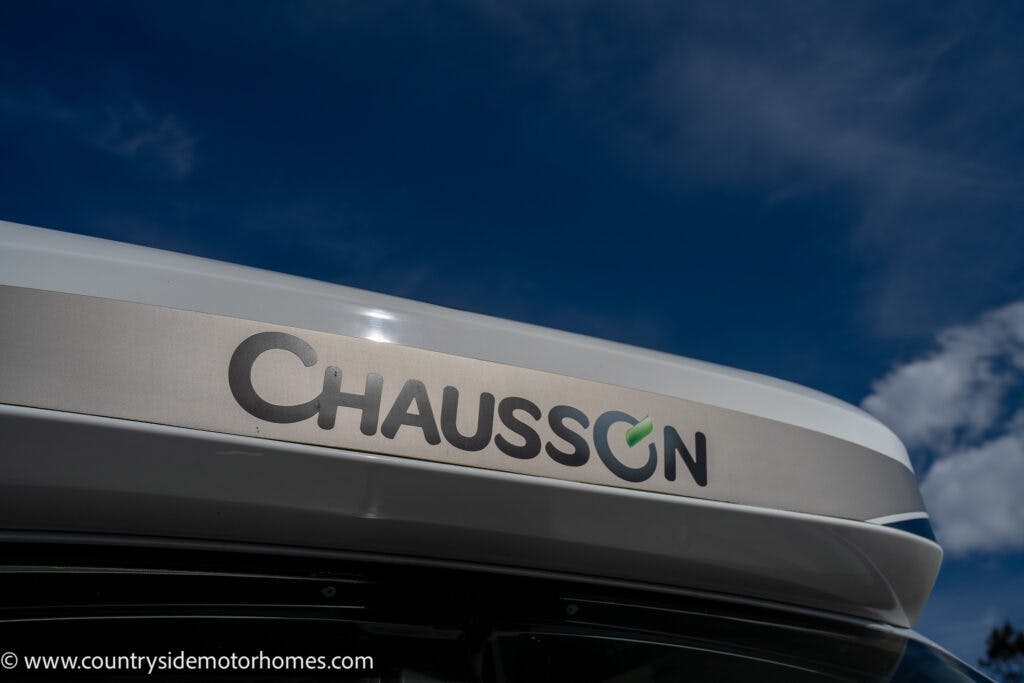 Close-up photo of the logo on a 2021 Chausson 778 Premium motorhome against a blue sky with a few clouds. The logo includes the brand name "Chausson" in black and a green-blue circle around the letter "O." A website URL is visible in the bottom left corner.