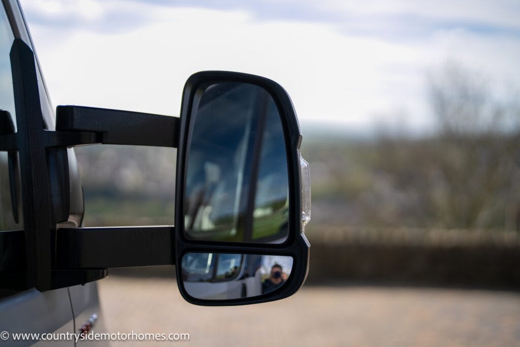 Close-up of a side mirror on a 2021 Chausson 778 Premium motorhome with a view of the landscape reflecting in the mirror. The background shows a blurred outdoor scene with trees and sky. The website "www.countrysidemotorhomes.com" is visible in the lower left corner.