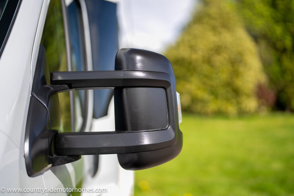 Close-up of a side mirror on a 2021 Chausson 778 Premium, with a blurred green grass area and trees in the background. The black mirror is mounted on a protruding arm, showcasing its extended design commonly seen on larger vehicles.
