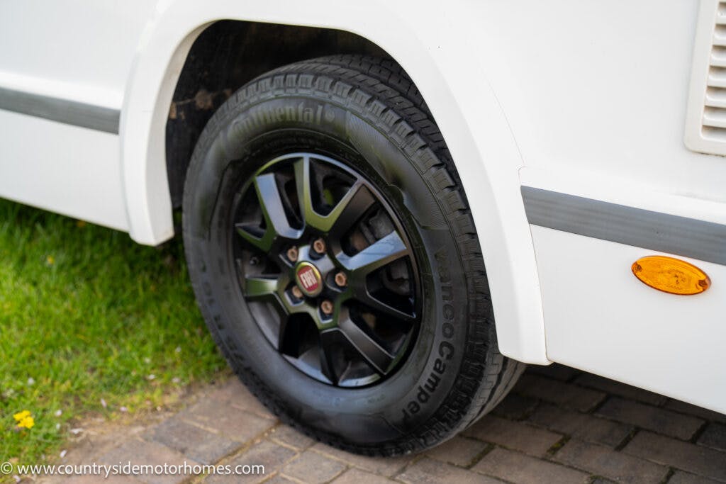 A close-up view of the rear wheel of a white 2021 Chausson 778 Premium motorhome. The tire is a Continental Vanco Camper model, mounted on a black alloy rim. The motorhome’s body side and part of a side marker light are visible. The vehicle is parked on a paved surface.