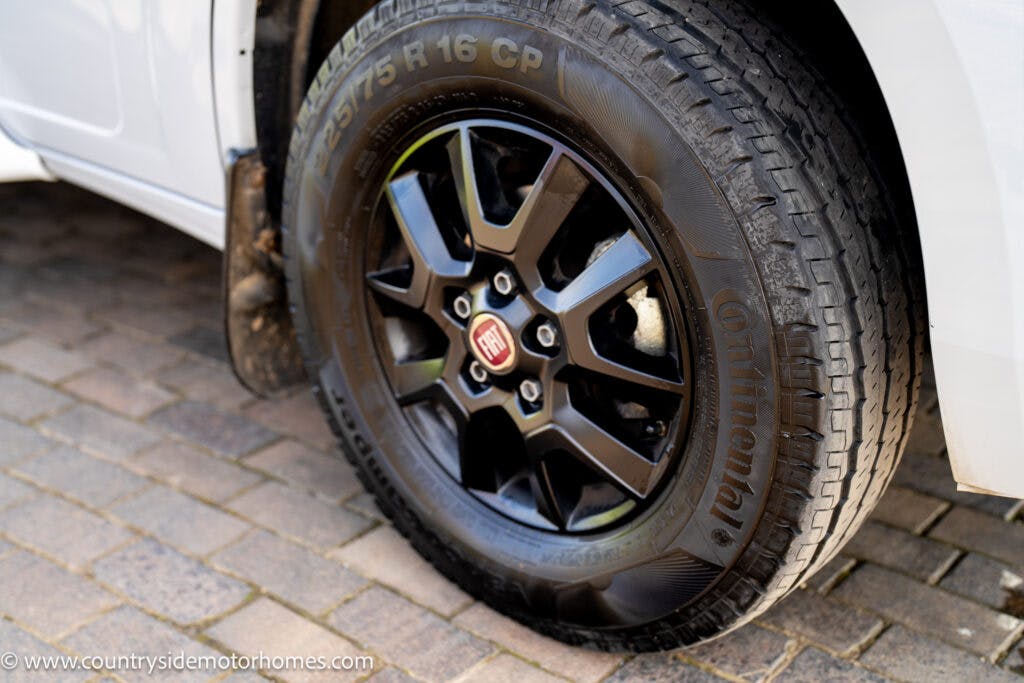 Close-up of a black Continental tire mounted on a 2021 Chausson 778 Premium vehicle. The tire is marked with the text "215/75 R 16 CP." The wheel features a black Fiat-branded hubcap. The vehicle is parked on a cobblestone surface. In the lower left corner, a watermark reads "www.countrysidemotorhomes.com".
