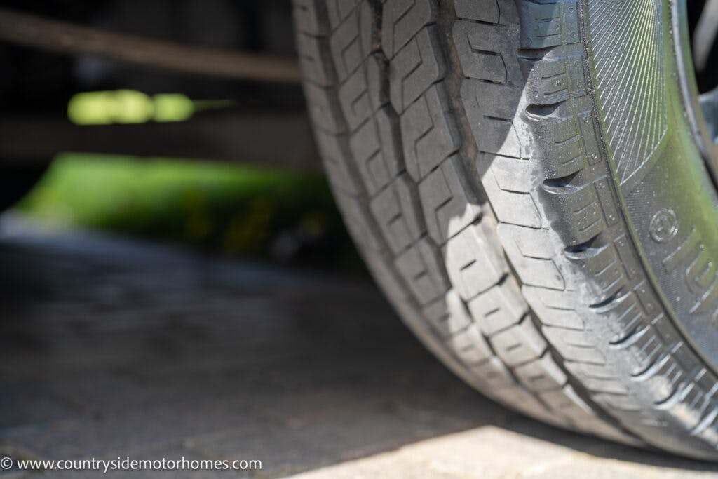 Close-up image of a tire with visible tread pattern mounted on a 2021 Chausson 778 Premium. Part of the wheel rim is visible on the right side, and the background shows a blurred outdoor setting, possibly a driveway.