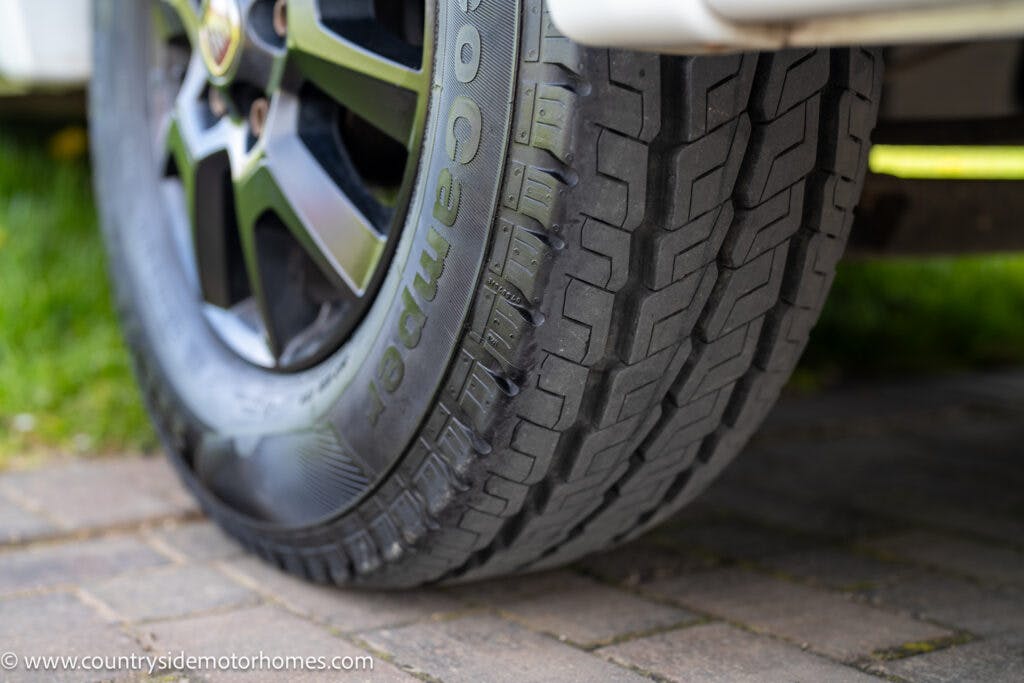 Close-up of a vehicle tire on a brick-paved surface. The tire, part of the 2021 Chausson 778 Premium, has visible tread patterns and the word "Cooper" on the sidewall. The surroundings include some grass and the bottom edge of the vehicle. A watermark reading "www.countrysidemotorhomes.com" is at the bottom left.