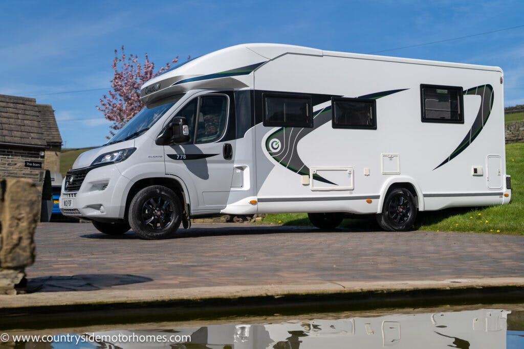 A 2021 Chausson 778 Premium motorhome with black and green accents is parked on a paved surface in front of a stone building. The motorhome features black wheels, multiple windows, and a storage compartment. The scene appears to be on a bright day with a clear blue sky.