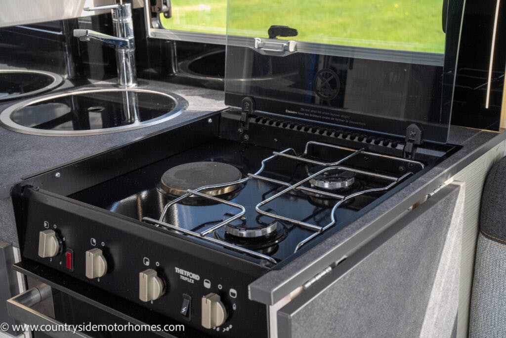 A compact kitchen inside the 2021 Chausson 778 Premium motorhome features a two-burner gas stove with a glass lid, control knobs below, and a round sink to the left. The stove is clean and housed in a modern countertop setting with a scenic outdoor view through the window.