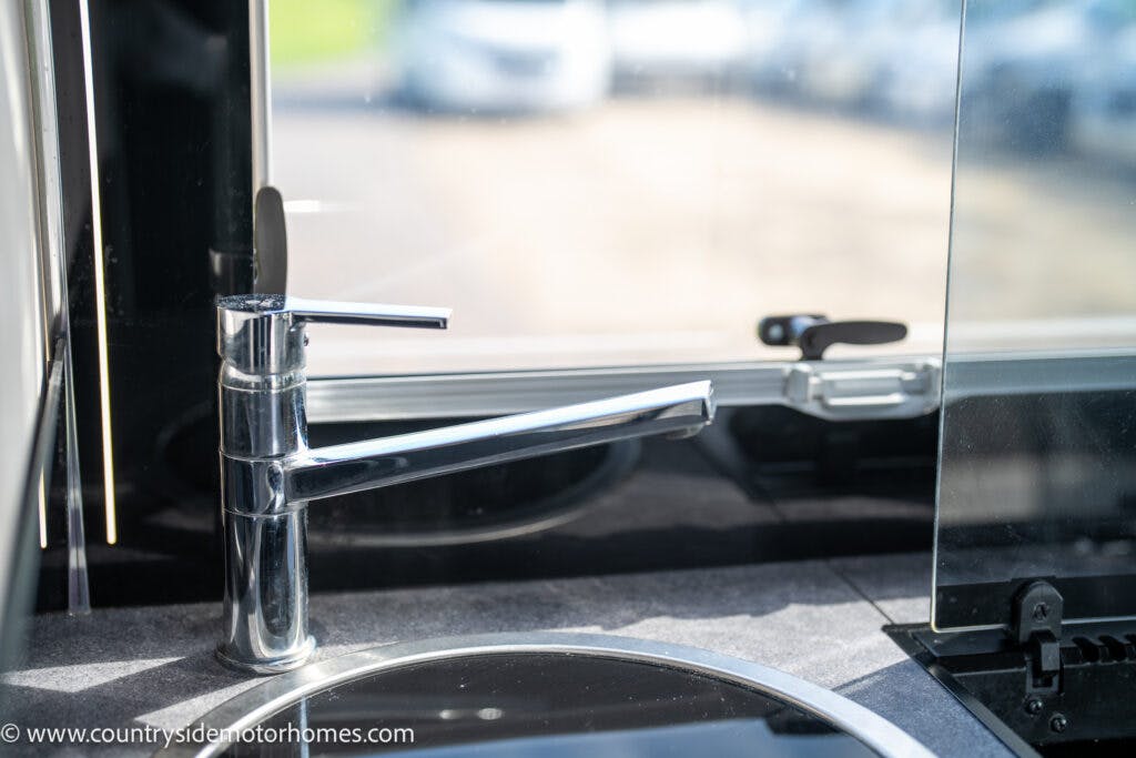 A close-up of a modern kitchen sink faucet installed in a 2021 Chausson 778 Premium motorhome. The faucet has a sleek, chrome finish and is positioned next to a round sink. Behind the faucet, a window provides a view of parked vehicles outside, while the dark countertop enhances its elegance.