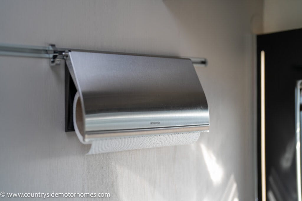 A paper towel holder is mounted on a wall, holding a roll of white paper towels. The holder, reminiscent of the modern design seen in the 2021 Chausson 778 Premium, has a silver metallic cover and is attached to a rail system with two small brackets. The wall background is light-colored and smooth.
