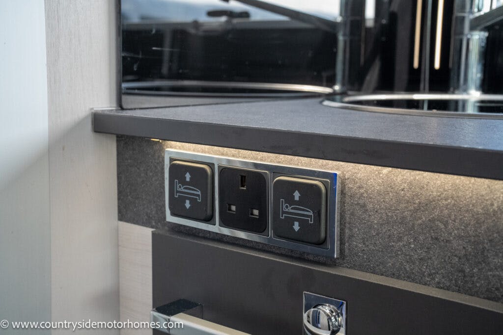 A close-up view of an electrical outlet panel in a 2021 Chausson 778 Premium motorhome. The panel features a standard electrical socket in the center and two adjacent switches with bed icons, indicating control of bed-related functions. A countertop and backsplash are visible above.