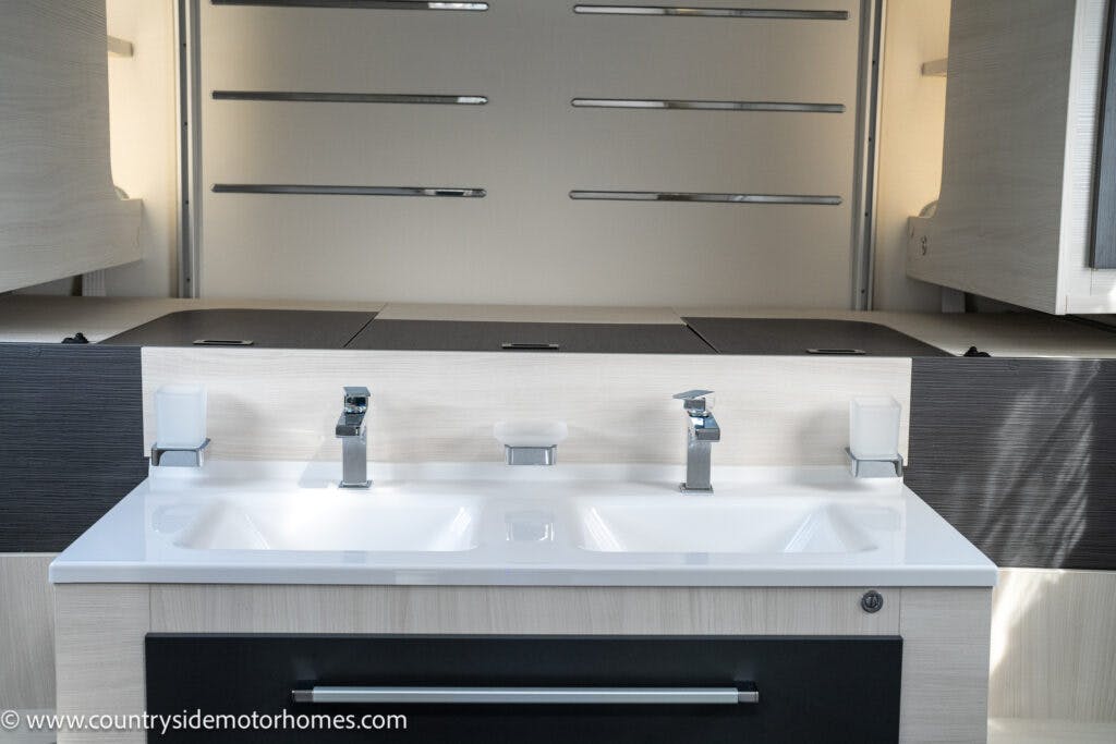 A modern bathroom vanity on the 2021 Chausson 778 Premium with a white double sink, two faucets, and silver handles. The countertop is surrounded by light wooden cabinets and drawers. Mirrored panels sit above the sinks with a website URL in the bottom left corner.