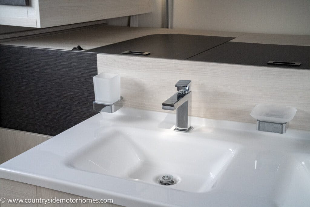 A modern bathroom sink in the 2021 Chausson 778 Premium with a rectangular basin, sleek metal faucet, and center drain. To the left of the faucet is a square soap dispenser, while to the right is a matching soap dish. The light-colored countertop features stylish dark accents.