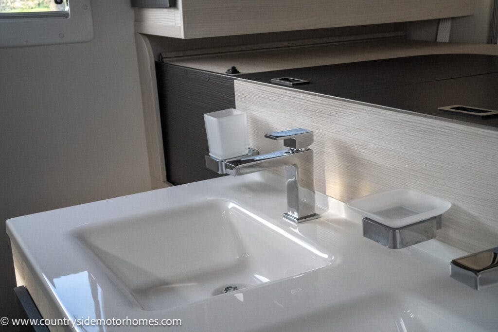 A modern bathroom sink with a rectangular basin, a chrome faucet, a white soap dispenser, and a soap dish. The background features light and dark wood paneling with a storage compartment. This elegant setup is part of the 2021 Chausson 778 Premium. Image credited to www.countrysidemotorhomes.com.