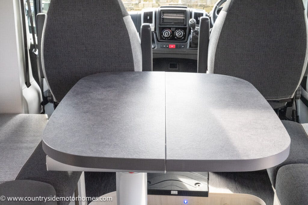 The image shows the dining area of a 2021 Chausson 778 Premium motorhome, featuring a grey table with a visible seam in the middle and two grey upholstered driver and passenger seats. The dashboard of the motorhome is also visible in the background.