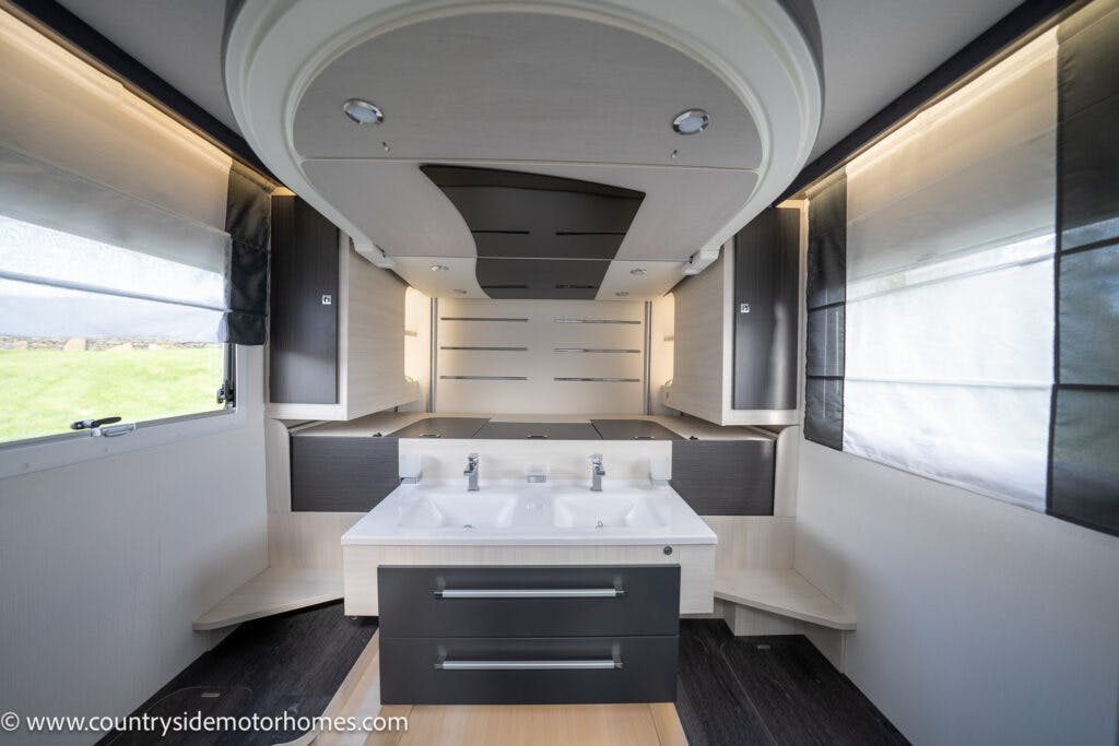 Interior view of a spacious and modern RV bathroom in the 2021 Chausson 778 Premium, featuring two sinks, a large mirror, and ample storage space. The decor is sleek with dark and light contrasting tones. Windows on both sides provide natural light. The URL countryside motor homes is visible.