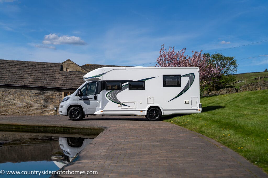 A white 2021 Chausson 778 Premium motorhome is parked on a paved driveway next to a rustic stone building. In the background, there are green fields and a flowering tree. The sky is clear with a few clouds.