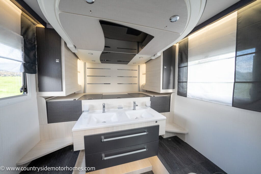 Interior view of the 2021 Chausson 778 Premium motorhome bathroom with a sleek, black and beige color scheme. It features a dual-sink vanity with two drawers, flanked by cabinets, and illuminated by overhead lighting. Blinds cover the windows on either side.