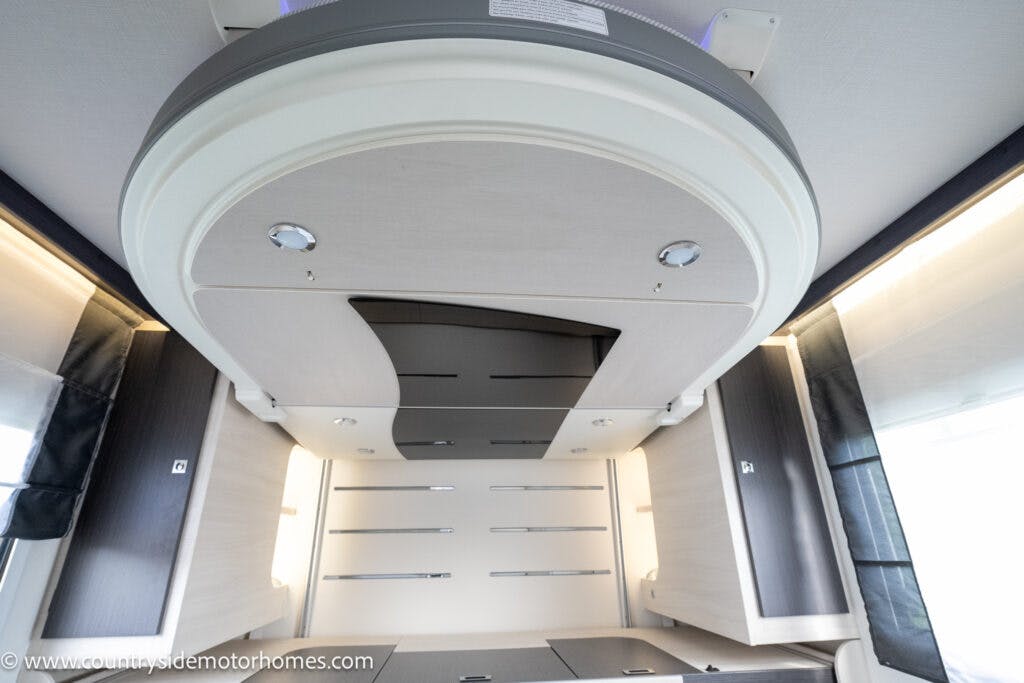 The interior of the 2021 Chausson 778 Premium motorhome boasts a modern design, featuring a curved, light-colored ceiling panel with integrated lighting. Surrounding cabinets have dark edges, and natural light streams through windows on either side, creating an inviting ambiance.