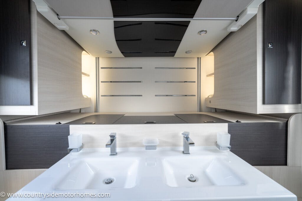 Modern motorhome bathroom interior in the 2021 Chausson 778 Premium features a white double sink with dual faucets, mirrors, and dark wood cabinets. Overhead lighting illuminates the room. Two soap dispensers are positioned on either side of the sinks.