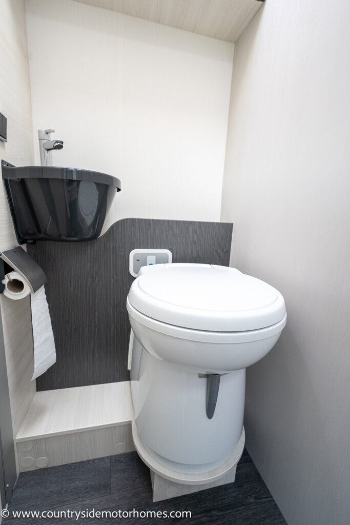 A compact bathroom in the 2021 Chausson 778 Premium motorhome, featuring a white toilet and a black sink with a chrome faucet mounted to the wall. The sink is above a small countertop, with a toilet paper holder on the left. The floor is dark gray, completing this sleek and modern space.