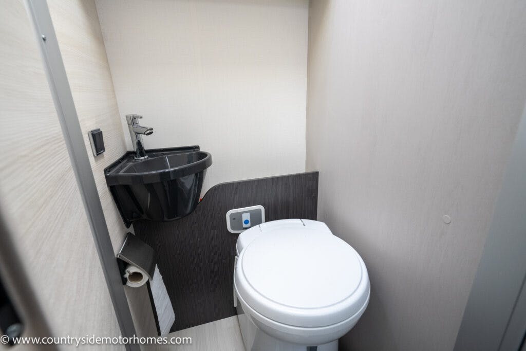 A compact bathroom in the 2021 Chausson 778 Premium RV features a white toilet with a small black sink above it and a toilet paper roll holder on the left wall. The light-colored walls and brushed metal faucet add a sleek touch. The space is neatly organized, maximizing functionality.