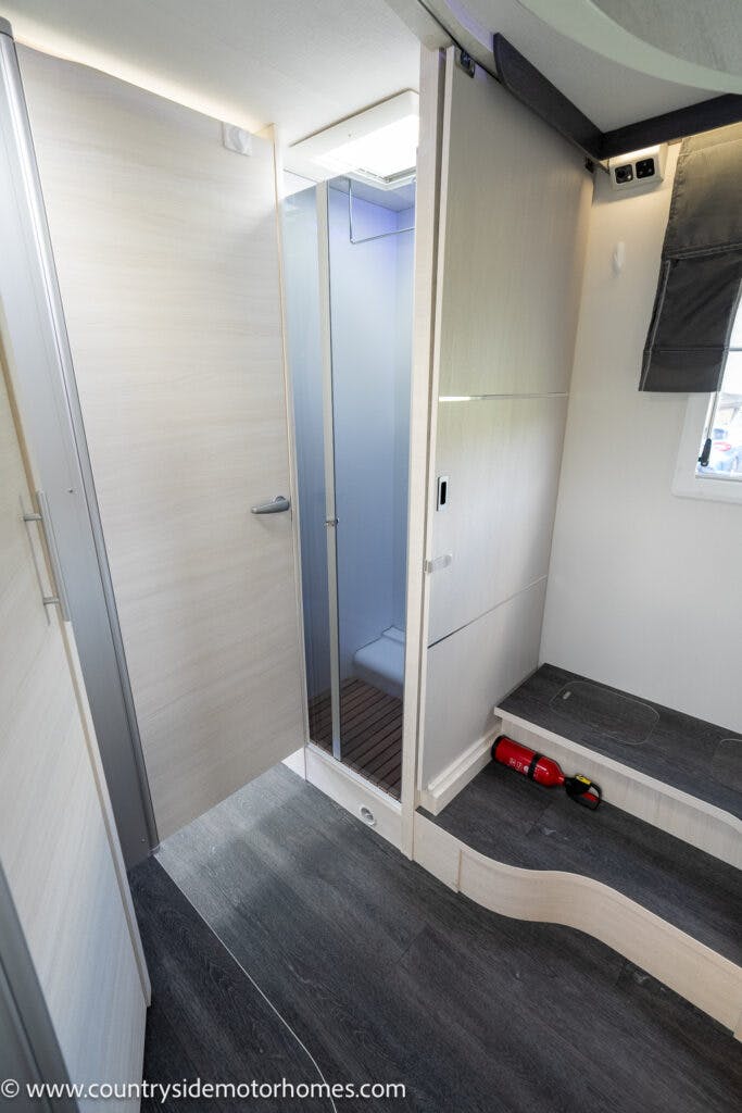 The image shows the interior of a 2021 Chausson 778 Premium motorhome featuring a hallway with wood-patterned walls and flooring. On the left is a sliding door to the bathroom, revealing a toilet and a shower. On the right is a bench with fire safety equipment on the step.