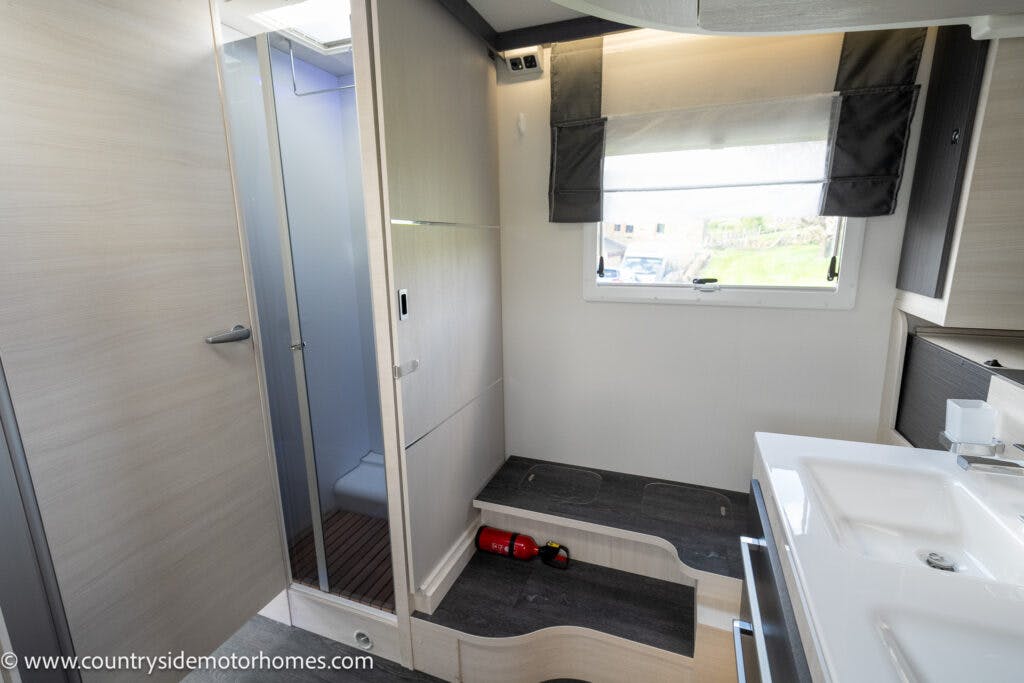 The 2021 Chausson 778 Premium motorhome interior features a shower area with a closed door on the left, a staircase leading to a raised sleeping area with two red wine bottles stored beneath a step, a window with a blind, and a sink with a mirror on the right.