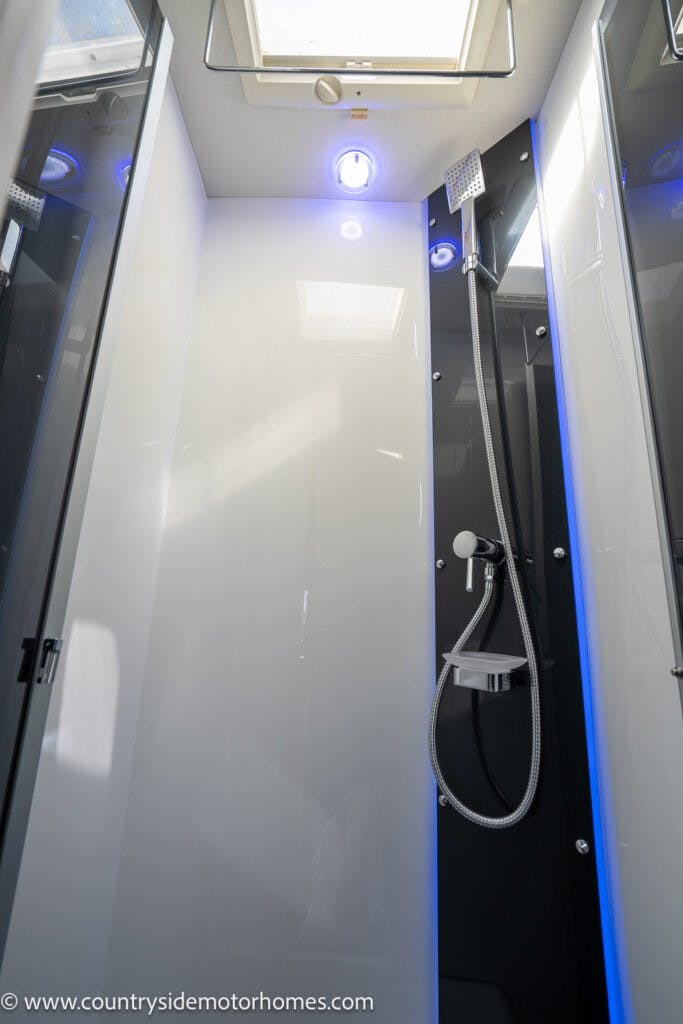 A modern shower area inside the 2021 Chausson 778 Premium motorhome, featuring a sleek white and black design. The showerhead is mounted on a sliding rail with a flexible hose. The space is illuminated by both natural light from a skylight and blue LED lights.