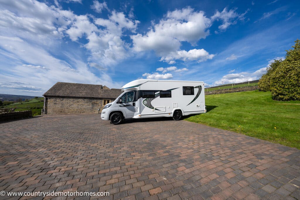 A 2021 Chausson 778 Premium motorhome is parked on a large paved driveway next to a stone building with a brown roof. The driveway is surrounded by a well-maintained lawn, and a blue sky with scattered clouds is visible in the background.