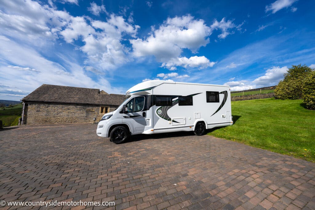 A white 2021 Chausson 778 Premium motorhome is parked on a paved area adjacent to a stone building. It features green and black graphics on its side. In the background, there's a grassy hill and a partly cloudy blue sky. The website www.countrysidemotorhomes.com is visible.