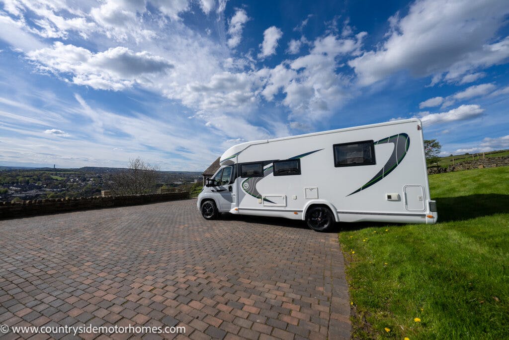 A white 2021 Chausson 778 Premium motorhome is parked on a cobblestone area next to a grassy hill under a blue sky with scattered clouds. In the distance, a town is visible. The motorhome features green and black detailing.