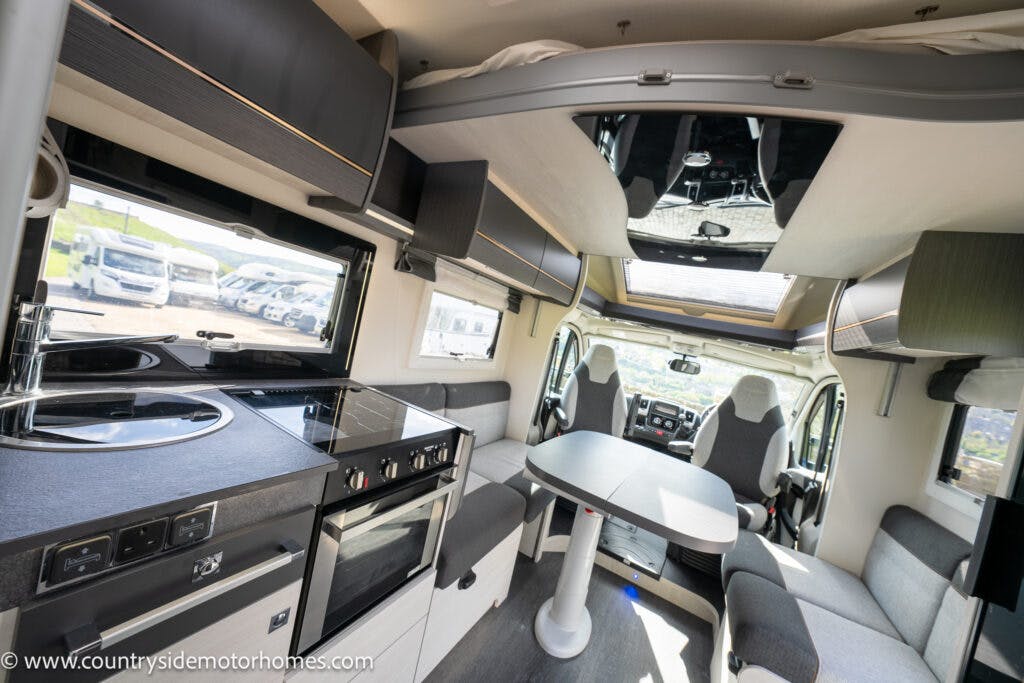 Interior view of the 2021 Chausson 778 Premium motorhome featuring a kitchen area with a stove and sink on the left, a dining table with seating in the center, and the driver and passenger seats in the front. There is ample natural light from the large windows and skylights.